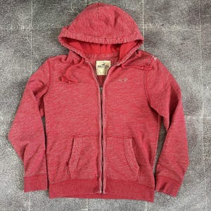 May include: Red zip-up hoodie with a drawstring hood and two front pockets. The hoodie features the Hollister logo on the chest and a tag on the inside collar. The fabric has a heathered appearance.