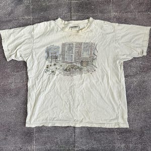 以下が含まれることがあります： クリーム色のTシャツ。犬、椅子、花のある素朴な小屋のシーンのグラフィックが特徴です。イラストは、グレー、ブラウン、グリーンの落ち着いた色調です。シャツは半袖で、クルーネックです。