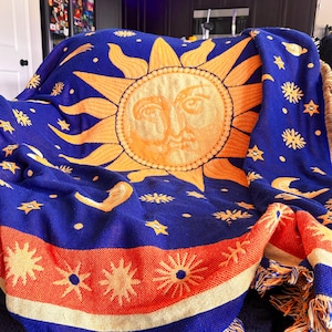 May include: A woven throw blanket featuring a large central sun with a face, surrounded by stars, moons, and comets on a deep blue background. The bottom edge has a wide orange band with sun motifs and a fringed border in orange and blue threads.