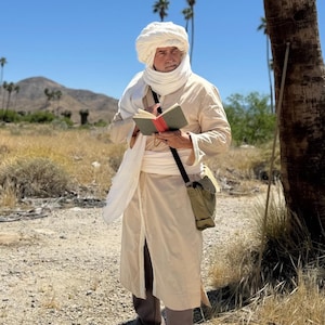 May include: A person in a cream-colored robe and turban, holding a small book with a red cover. They wear a white scarf and carry a green messenger bag. The background features a desert landscape with palm trees and mountains under a clear blue sky.