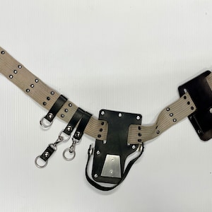 May include: A tan canvas work belt with black leather accents and a black leather tool holder. The belt has multiple metal loops and a snap closure.