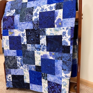 May include: A patchwork quilt in shades of blue, displayed on a wooden rack. The quilt features various patterns, including floral, polka dots, and abstract designs. The quilt is made of squares and rectangles of different blue fabrics.