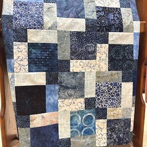 May include: A patchwork quilt with a variety of blue fabrics, including solid, patterned, and textured squares. The quilt is draped over a wooden rack, showcasing the different shades and designs.