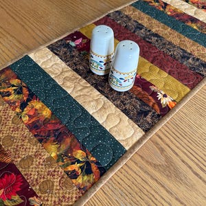 May include: A quilted table runner with a patchwork design featuring various autumn-themed fabrics in shades of brown, green, and gold. Two white ceramic salt and pepper shakers with floral patterns sit on top of the runner.