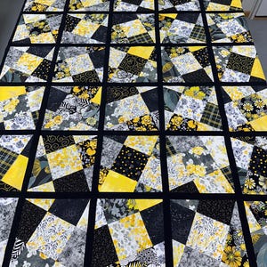 May include: A patchwork quilt with a geometric design. The quilt features a grid of squares, each divided into triangles of black, gray, yellow, and floral patterns. The quilt is bordered with a solid black fabric.