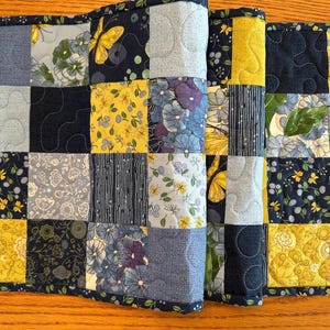 May include: A quilted table runner with a patchwork design featuring blue, yellow, and white floral patterns. The runner is made of fabric squares sewn together to create a unique and decorative piece.
