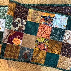 May include: A patchwork table runner with a fall theme. The runner is made of various fabrics with autumnal colors and patterns, including leaves, acorns, and branches. The runner has a green border.