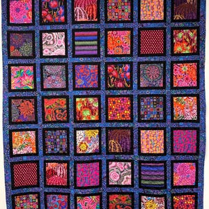 May include: A quilt featuring a grid of thirty-six square fabric patches in vibrant floral, geometric, and abstract patterns. Each square is framed by a solid black border, and the entire grid is separated by a blue patterned fabric lattice.