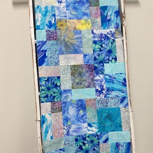 May include: A handmade quilt with a patchwork design, featuring various shades of blue, teal, and purple. The quilt is displayed on a white wooden ladder. The fabric patterns include floral prints and abstract designs.