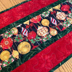 Puede incluir: Un camino de mesa acolchado con un panel central sobre fondo negro decorado con adornos navideños rojos, dorados y blancos y hojas de acebo verdes, bordeado por franjas verde oscuro y paneles exteriores rojos con patrones cosidos.
