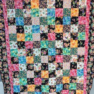 May include: A colorful quilt with a patchwork design featuring hummingbirds and floral patterns. The quilt is made up of squares of different colors and patterns, including pink, blue, green, yellow, and orange. The quilt is edged with a black border.
