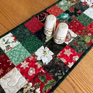 May include: A Christmas-themed table runner with a patchwork design featuring red, green, and white squares. The runner has a dark green border and two white salt and pepper shakers with decorative bands.