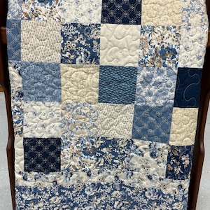 May include: A patchwork quilt in shades of blue, cream, and tan, draped over a wooden chair. The quilt features various floral and geometric patterns in square blocks. The border has a floral design. The quilt is a cozy home decor item.