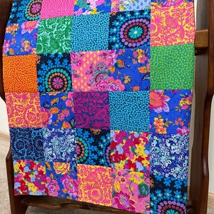 May include: A colorful patchwork quilt with a variety of floral and geometric patterns in shades of pink, blue, green, and yellow. The quilt is displayed on a wooden rack, showcasing its vibrant design and intricate details. The quilt's dimensions are not visible.