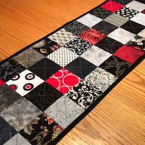 May include: A black and white patchwork table runner with red squares. The runner is made of different fabrics with various patterns, including polka dots, floral designs, and geometric shapes.