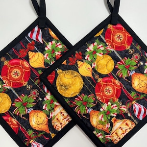 May include: Two square quilted pot holders featuring a black background with a repeating pattern of red and gold Christmas ornaments, green holly leaves, and red berries. Each pot holder has a solid black fabric border and a black hanging loop.