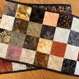 May include: A quilted table runner with a patchwork design featuring various shades of brown, beige, and cream. The runner has a black border and is made of fabric squares with different patterns.
