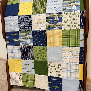 May include: A patchwork quilt with a lake theme, featuring squares of various patterns and colors. The quilt includes images of sailboats, canoes, trees, and text that reads "AT THE LAKE" and "The Best Days Are". The quilt is draped over a wooden bench.