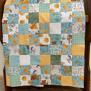 May include: A patchwork quilt featuring a repeating pattern of safari animals including lions, zebras, elephants, and leopards. The fabric squares alternate between yellow, light blue, and green, with text prints that read Be Brave, Be Proud, and Love.