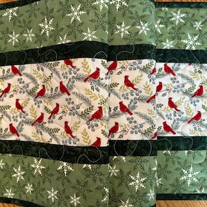 May include: A quilted table runner featuring a repeating pattern of red cardinals perched on branches with green leaves. The runner has green snowflake and dark green patterned borders. The dimensions are approximately 14 inches wide and 36 inches long.