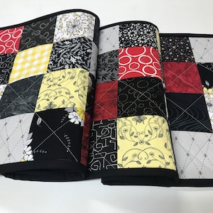 May include: A black and white quilted table runner with red and yellow accents. The runner is made of patchwork fabric with different patterns and designs. It has a black border and is quilted with a diamond pattern.
