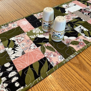 May include: A floral patchwork table runner with a green border, featuring pink, white, and black floral patterns. Two white salt shakers with decorative bands sit atop the runner. The runner is on a wooden table.