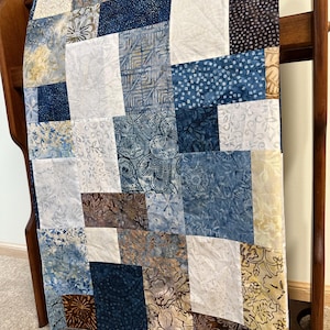 May include: A patchwork quilt featuring a variety of rectangular fabric blocks in shades of navy blue, light blue, white, and brown. The fabric patterns include floral motifs, geometric shapes, and batik-style designs arranged in a grid layout.