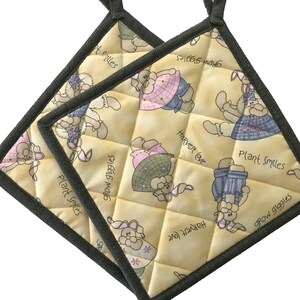 May include: Two square quilted pot holders with a light yellow background. The pot holders feature a repeating pattern of cartoon bunnies in various outfits. Text includes "Plant Smiles", "Grow Giggles", and "Harvest Love".