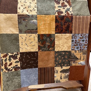May include: A patchwork quilt with a variety of earth-toned fabrics, including brown, beige, and green squares. Some squares feature nature-themed patterns like leaves, ducks, and cattails. The quilt is draped over a wooden chair.