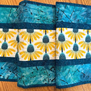 May include: A quilted table runner with a floral design. The runner features a repeating pattern of yellow and blue flowers on a white background, framed by teal and blue batik fabric. The runner is approximately 15 inches wide.