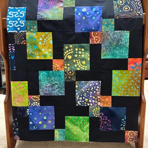 May include: A patchwork quilt featuring a grid of solid black fabric squares alternating with colorful batik fabric squares. The batik squares display various patterns including dots, swirls, and floral motifs in shades of blue, green, orange, and purple.