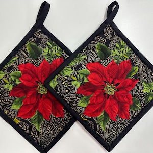 May include: Two square quilted pot holders featuring a vibrant red poinsettia floral pattern on a black background with intricate gold paisley details. Each pot holder is finished with a solid black fabric border and a small hanging loop.