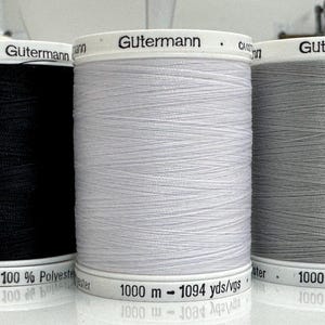 May include: Three spools of Gütermann 100% polyester sewing thread in black, white, and gray. Each spool features a white top and bottom cap with the brand name and a length of 1000 meters or 1094 yards printed clearly on the base of the spool.
