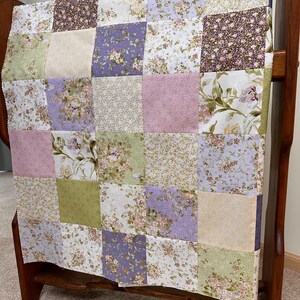 May include: A patchwork quilt with a floral pattern in shades of lavender, pink, green, and cream. The quilt is displayed on a wooden rack, showcasing its square blocks of various floral fabrics. The quilt's design is a mix of small and large floral prints.