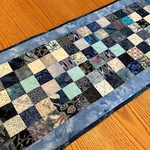 May include: A blue and white patchwork table runner with a blue border. The runner is made of various fabrics with different patterns and textures.