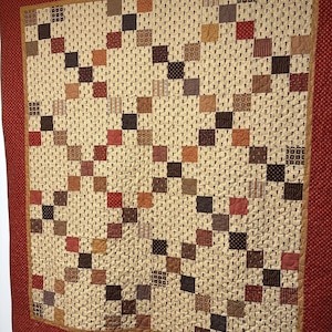 May include: A handmade quilt with a repeating pattern of squares in shades of brown, rust, and tan. The squares are arranged in a grid-like design against a cream-colored background. The quilt is framed by a solid rust-colored border.