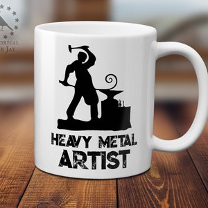 May include: White ceramic mug with a black silhouette of a blacksmith working at an anvil. The text "Heavy Metal Artist" is printed below the image.