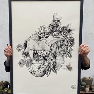 Peut inclure: Illustration en noir et blanc d'un crâne d'animal avec des fleurs et une abeille. Le crâne est entouré de fleurs et de feuilles, et une abeille vole dans le coin supérieur droit. L'illustration est réalisée dans un style détaillé et réaliste.