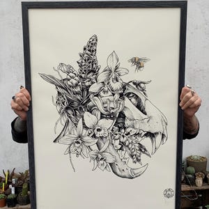 Peut inclure: Illustration en noir et blanc d'un crâne avec des fleurs et une abeille. Le crâne est entouré de fleurs, dont des orchidées et du gingembre. L'abeille vole au-dessus du crâne.