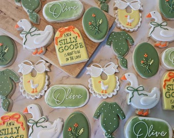 May include: A collection of decorated sugar cookies featuring white geese with green bows, yellow baby bibs with white geese, green baby onesies with white dots, oval cookies with floral designs, and rectangular cookies with the text A NEW SILLY GOOSE IS ABOUT TO BE ON THE LOOSE.