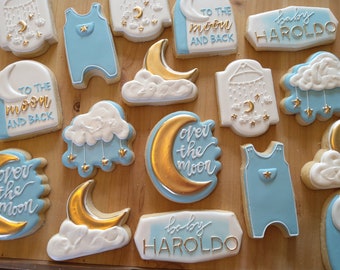 May include: A set of 12 decorated sugar cookies with a blue and gold color scheme. The cookies feature a variety of designs including a baby onesie, a crescent moon, a cloud, and the text "To the moon and back" and "Baby Haroldo".