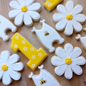 May include: A set of nine decorated sugar cookies featuring a yellow number one, white daisies with yellow centers, and white bee-shaped cookies with yellow and black accents.