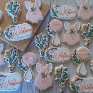 Puede incluir: Una colecci&oacute;n de galletas de az&uacute;car decoradas con un tema floral con el texto "A Little Wildflower is on her way!" en letras rosa y naranja. Las galletas est&aacute;n decoradas con glaseado rosa, blanco y verde y presentan dise&ntilde;os florales, sonajeros para beb&eacute;s y monos para beb&eacute;s.