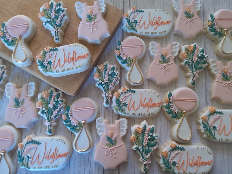 Puede incluir: Una colecci&oacute;n de galletas de az&uacute;car decoradas con un tema floral con el texto "A Little Wildflower is on her way!" en letras rosa y naranja. Las galletas est&aacute;n decoradas con glaseado rosa, blanco y verde y presentan dise&ntilde;os florales, sonajeros para beb&eacute;s y monos para beb&eacute;s.