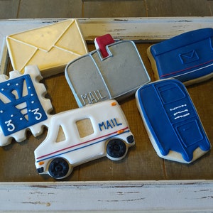 May include: A set of six decorated sugar cookies in the shape of mail-related items. The cookies are decorated with white, blue, red, and gray icing. The cookies include a mail truck, a mailbox, a stamp, and an envelope. The cookies are arranged in a white frame.