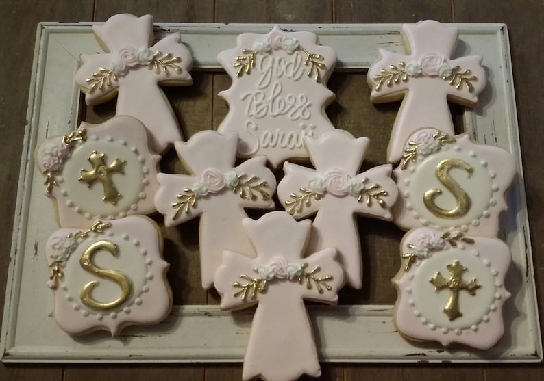 May include: Nine pink and gold decorated sugar cookies with a cross design. The cookies have gold lettering that says "God Bless Sarai" and the letter "S".