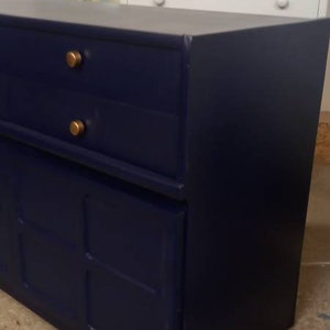 May include: A dark blue dresser with two drawers and a cabinet door. The dresser has gold knobs and is sitting on a concrete floor.