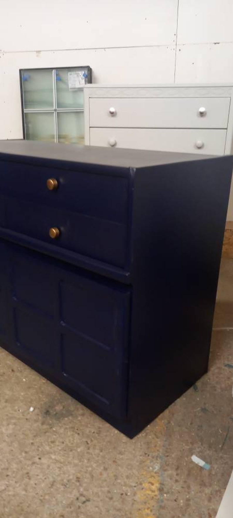 May include: A dark blue dresser with two drawers and a cabinet door. The dresser has gold knobs and is sitting on a concrete floor.