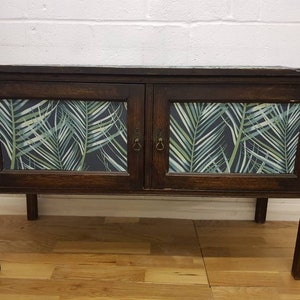 May include: A dark wood cabinet with two doors. The doors are decorated with a tropical leaf pattern wallpaper. The cabinet has a simple design and is sitting on a wooden floor.