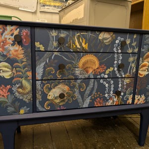 Op de afbeelding: Een donkerblauwe dressoir met drie lades en twee deuren. De lades en deuren zijn versierd met een bloemmotief met zeeleven, waaronder koraal, vissen en schelpen. De dressoir heeft zwarte metalen knoppen.